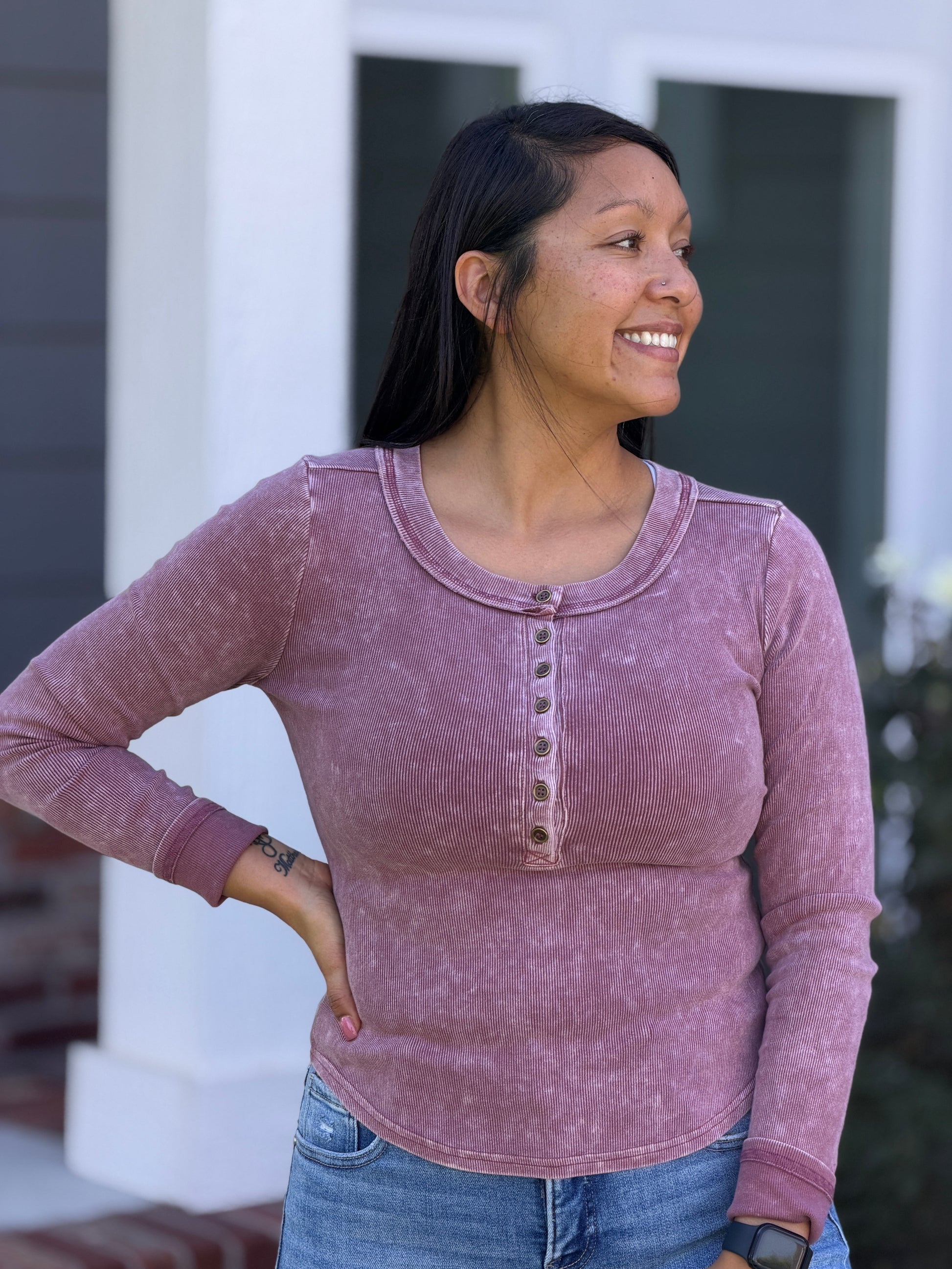 A woman wearing a dusty rose long-sleeve henley top with bronze buttons, paired with blue jeans, standing outdoors in front of a white house column.