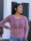 A woman wearing a dusty rose long-sleeve henley top with bronze buttons, paired with blue jeans, standing outdoors in front of a white house column.