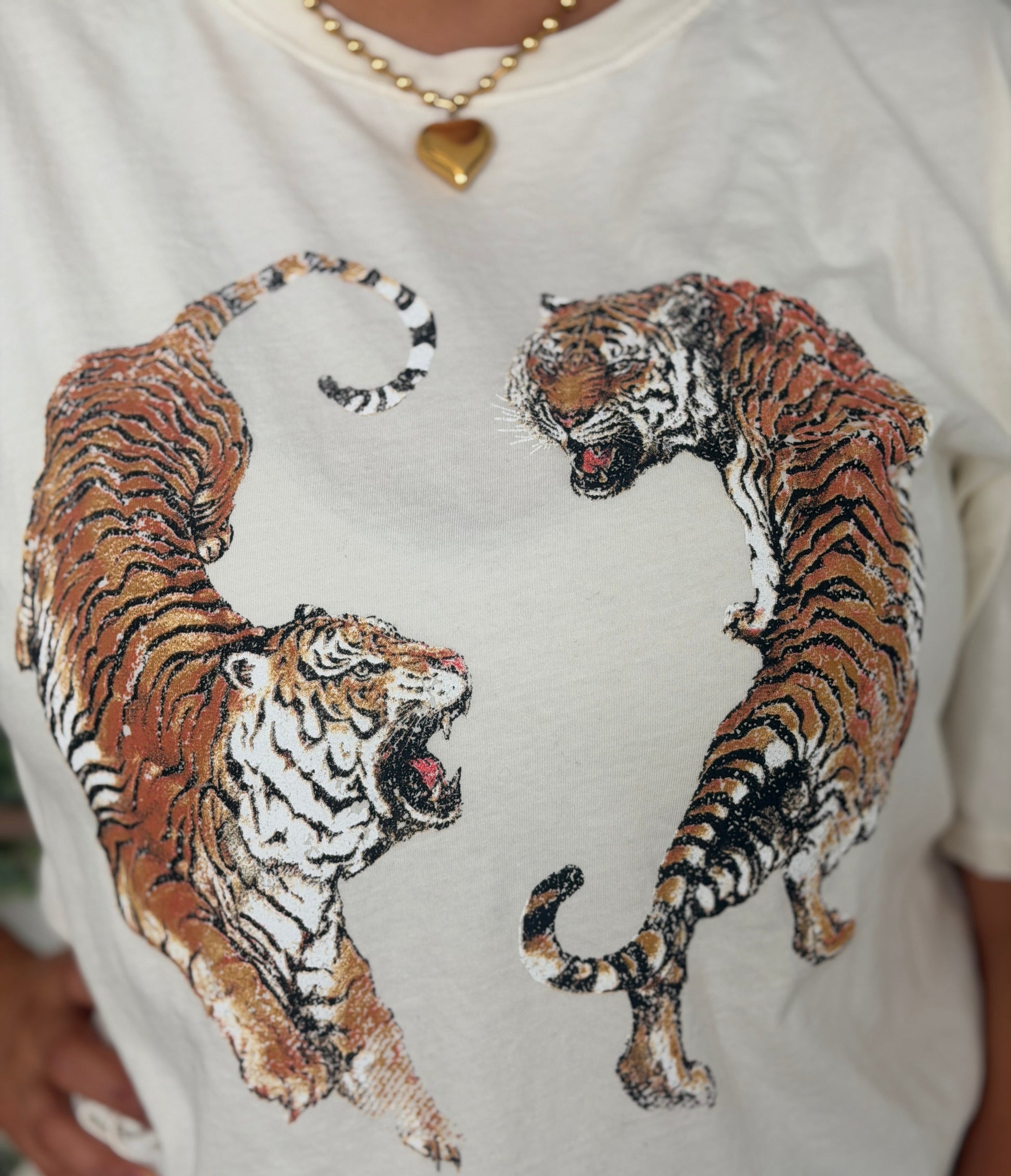 A close-up of a person wearing a white t-shirt with a graphic print of two roaring tigers facing each other. The person is also wearing a gold heart pendant necklace.