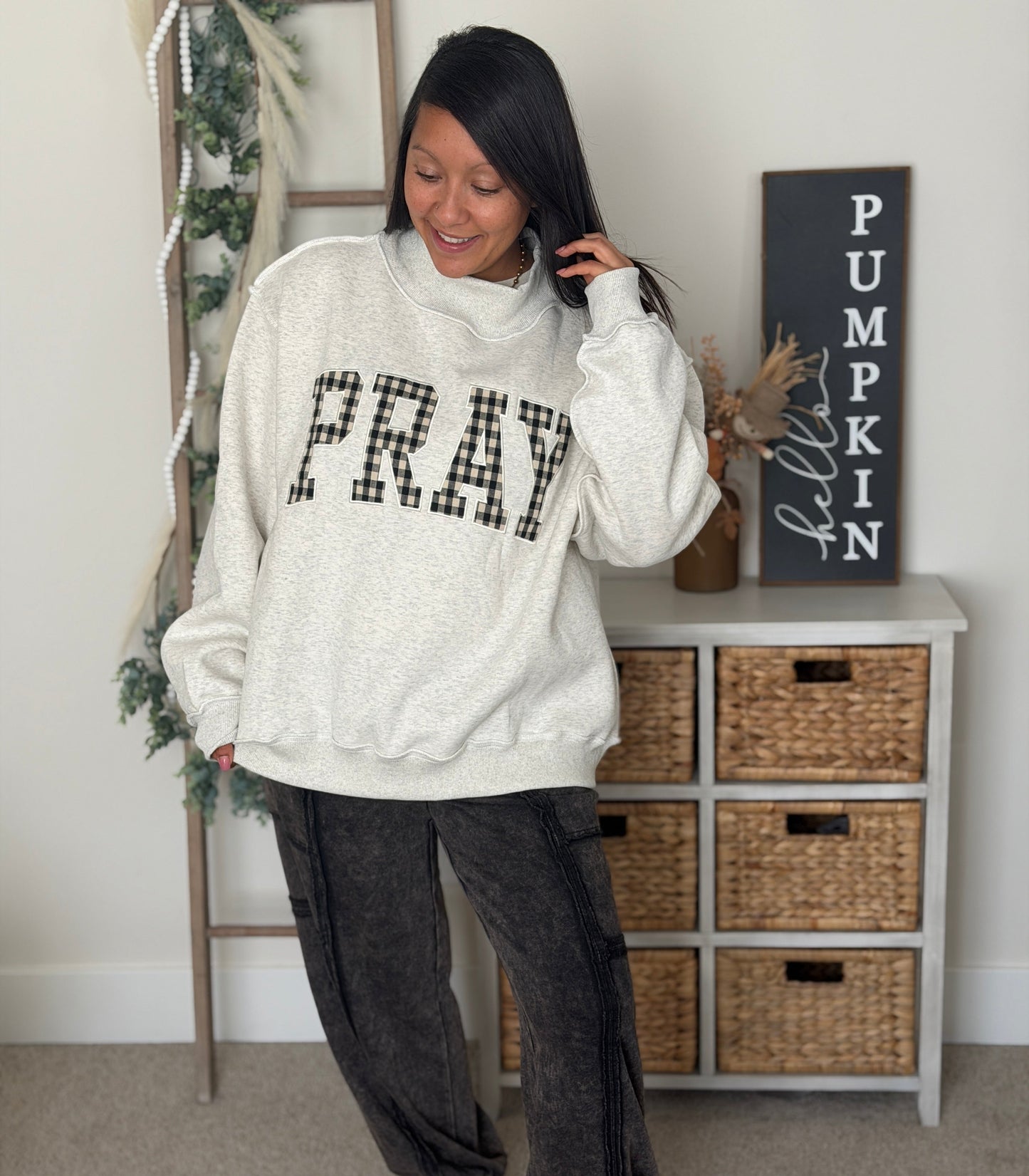 A woman wearing a cream-colored crewneck sweatshirt with 'PRAY' in black plaid letters, paired with dark gray relaxed-fit pants and white sneakers, standing in a decorated room.