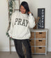 A woman wearing a cream-colored crewneck sweatshirt with 'PRAY' in black plaid letters, paired with dark gray relaxed-fit pants and white sneakers, standing in a decorated room.