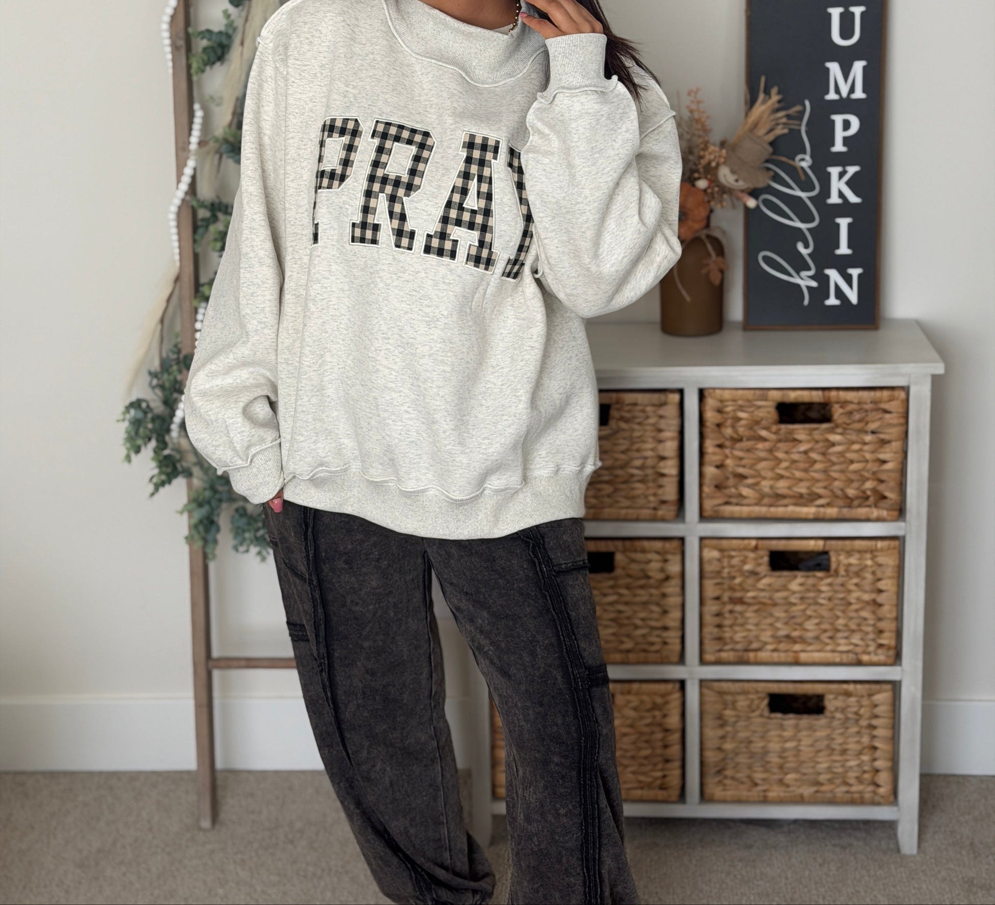 A woman wearing a cream heathered sweatshirt with 'PRAY' in black plaid letters, paired with dark gray cargo pants and white sneakers, standing in a decorated room next to a ladder and a dresser.