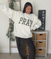 A woman modeling an oversized light gray sweatshirt with 'PRAY' in black plaid block letters, paired with dark gray jogger pants and white sneakers, standing in a decorated room with a ladder, wreath, and a 'PUMPKIN' sign.