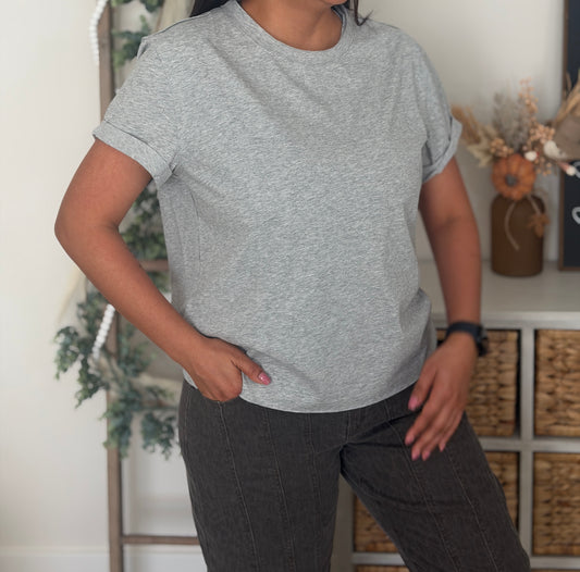 A person wearing a light heather gray short-sleeve crewneck t-shirt and dark gray pants, standing in a styled indoor room with shelves and decorative items in the background.