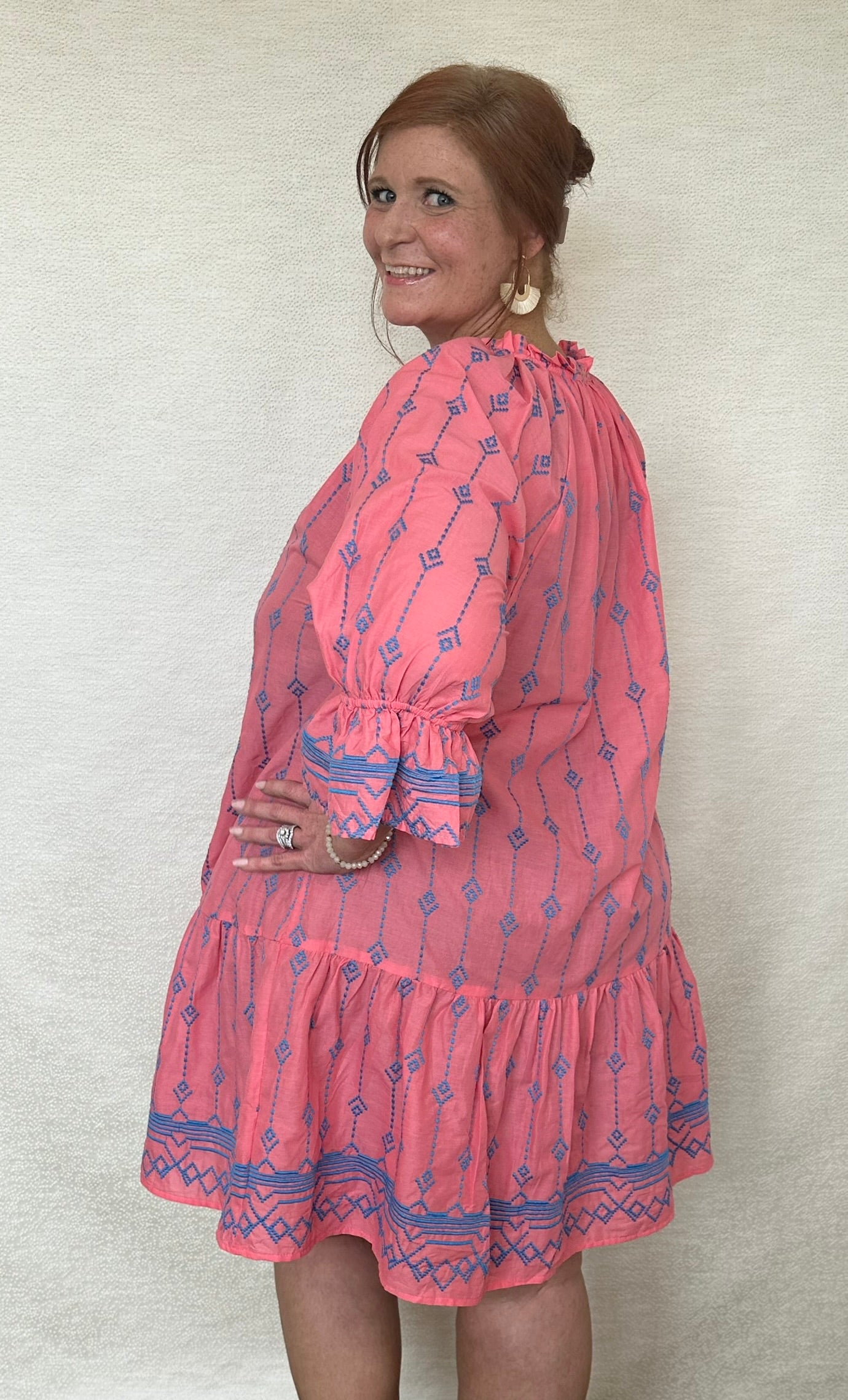 A woman wearing a pink tiered dress with blue embroidery, ruffled neckline, and tassel ties.
